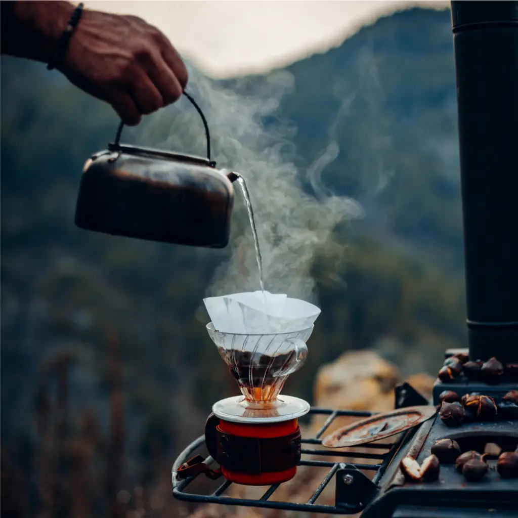 Une personne verse de l'eau chaude d'une bouilloire à travers un filtre à café dans une tasse, posée sur un réchaud portable, avec un paysage montagneux boisé en arrière-plan.