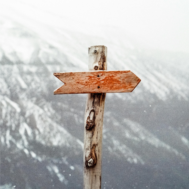 An old wooden signpost with a single weathered arrow pointing left, against a blurred background of a snowy mountainside.