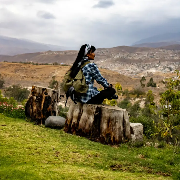 Une femme est assise sur une grande souche d’arbre surplombant une vallée, avec un ciel nuageux au-dessus et des collines sèches et vallonnées au loin. Elle a un sac à dos et porte une chemise à carreaux, préparée pour sa randonnée.