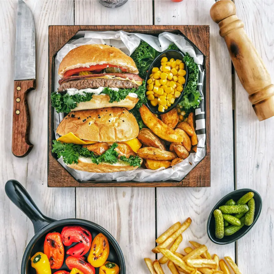 Un plateau en bois contient deux hamburgers avec de la laitue et des tomates, un petit plat de maïs et une portion de quartiers de pommes de terre. Autour du plateau se trouvent un couteau, un moulin à poivre, une poêle avec un mélange de poivrons, des frites et un petit plat de cornichons sur une table en bois clair.
