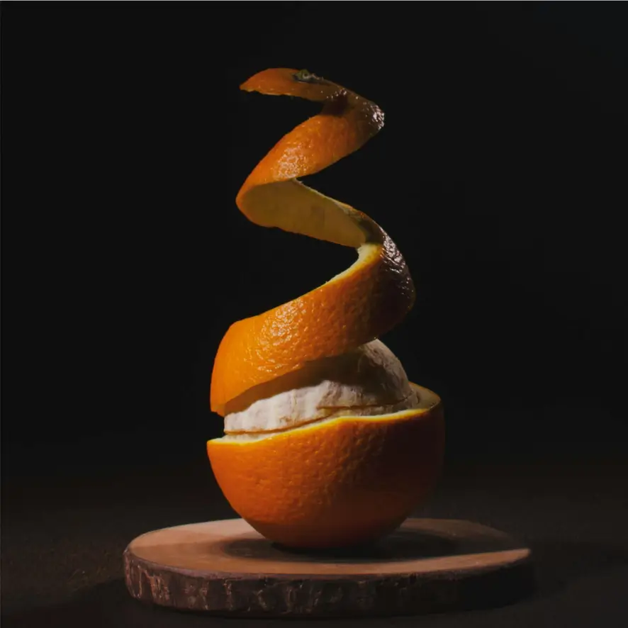  Une orange partiellement pelée repose sur un dessous de verre rond en bois sur un fond sombre. La peau s'enroule vers le haut au-dessus du fruit, mettant en valeur la moelle blanche et les segments juteux à l'intérieur. L'éclairage accentue la texture et la couleur vibrante de l'orange.
