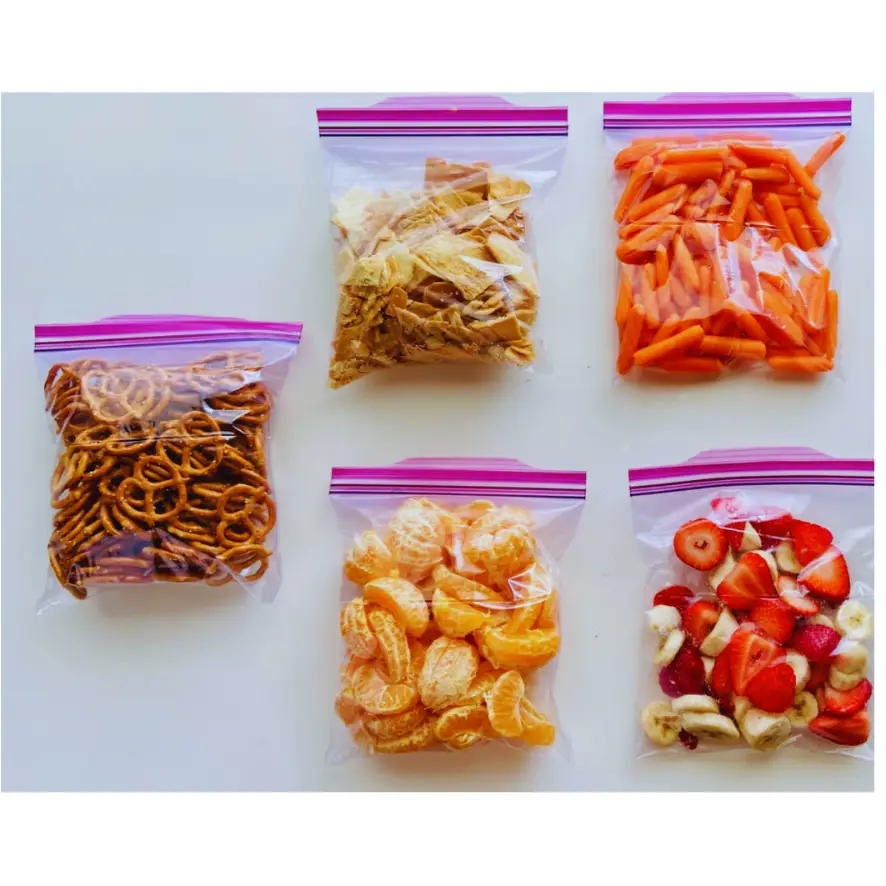 Five plastic zipper bags containing various snacks arranged on a white surface. The bags contain pretzels, dried apple slices, baby carrots, peeled tangerine segments and a mix of strawberry and banana slices.