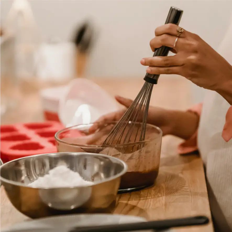 A person whisking chocolate batter in a glass bowl on a wooden counter. Nearby there is a stainless steel bowl with white powder and a red silicone mold. Other kitchen utensils and containers are blurred in the background.