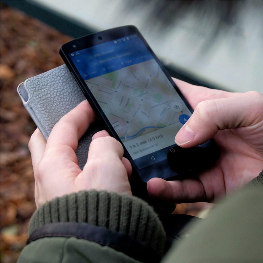 A person holds a smartphone displaying a map application suggesting navigation. The phone is in their right hand, with the left hand touching the screen. They are wearing a green jacket and holding a gray textured wallet or phone case.