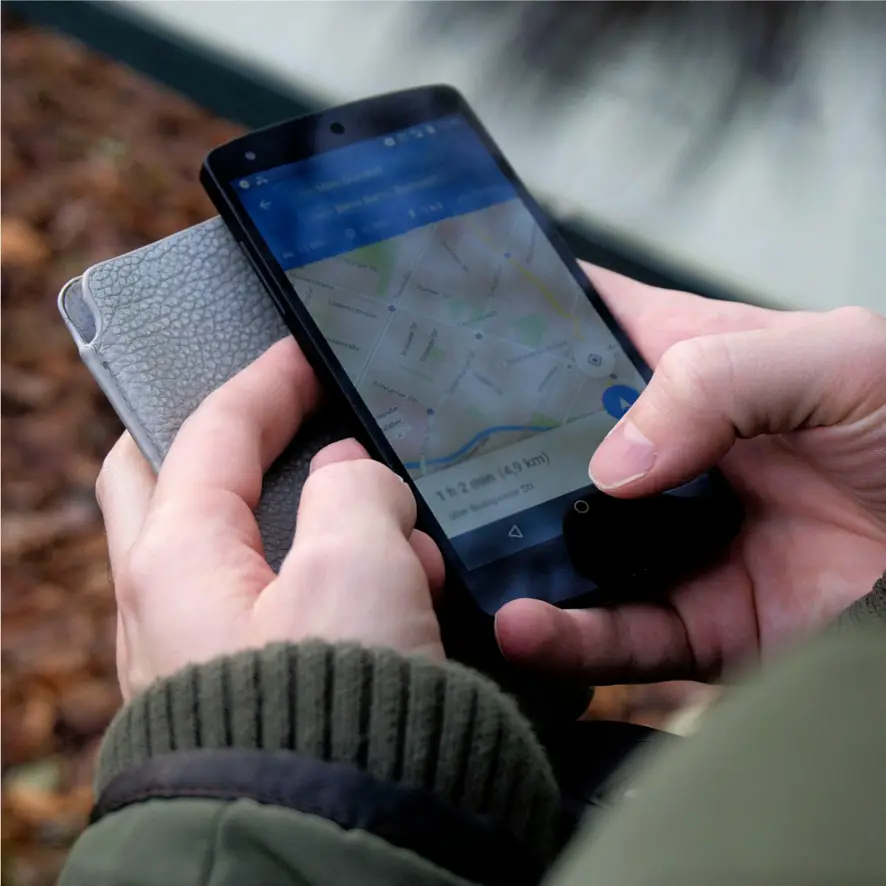 Une personne tient un smartphone affichant une application cartographique suggérant la navigation. Le téléphone est dans leur main droite, la main gauche touchant l’écran. Ils portent une veste verte et tiennent un portefeuille ou un étui de téléphone texturé gris.
