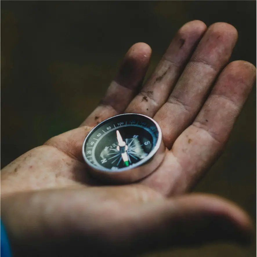 A dirty hand holds a small silver compass. The compass needle points north and the letters indicating the cardinal directions are visible. The background is blurred, suggesting an outdoor or nature setting.