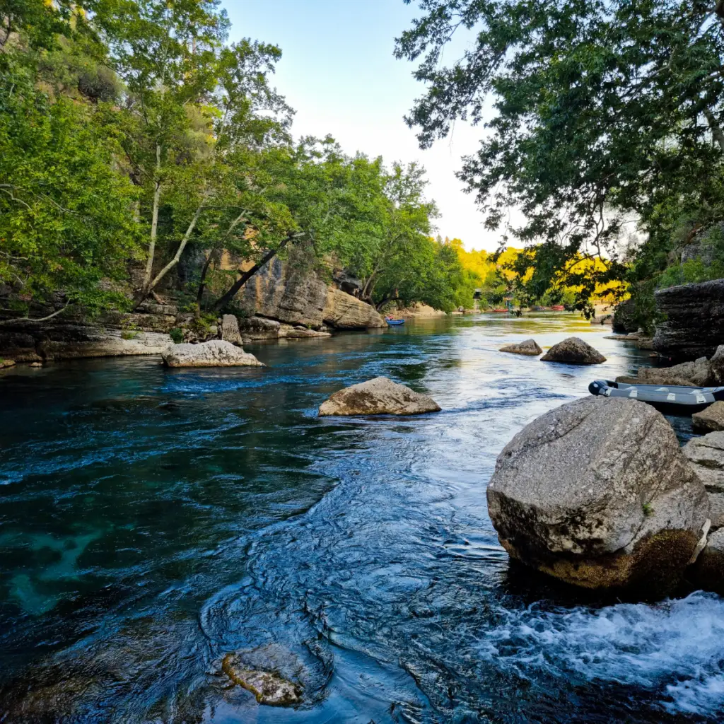 A serene river flows through a rocky landscape surrounded by green trees. Large rocks are scattered in the clear blue water and a small boat is moored along the right bank. The sky is clear, with the evening sun casting a warm glow.