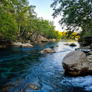 A serene river flows through a rocky landscape surrounded by green trees. Large rocks are scattered in the clear blue water and a small boat is moored along the right bank. The sky is clear, with the evening sun casting a warm glow.