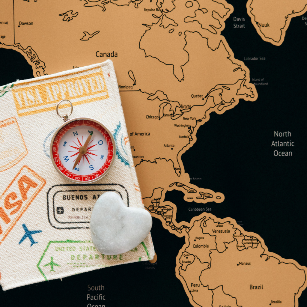 A fabric-covered notebook with travel-related stamps, a small red and white compass, and a heart-shaped stone placed on top. In the background is a map highlighting North and South America with the surrounding oceans labeled.
