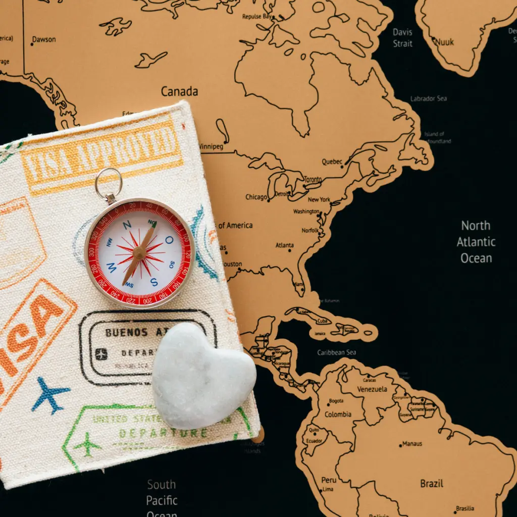 Un carnet recouvert de tissu avec des timbres liés au voyage, une petite boussole rouge et blanche et une pierre en forme de cœur posée dessus. En arrière-plan se trouve une carte mettant en évidence l’Amérique du Nord et l’Amérique du Sud avec les océans environnants étiquetés.
