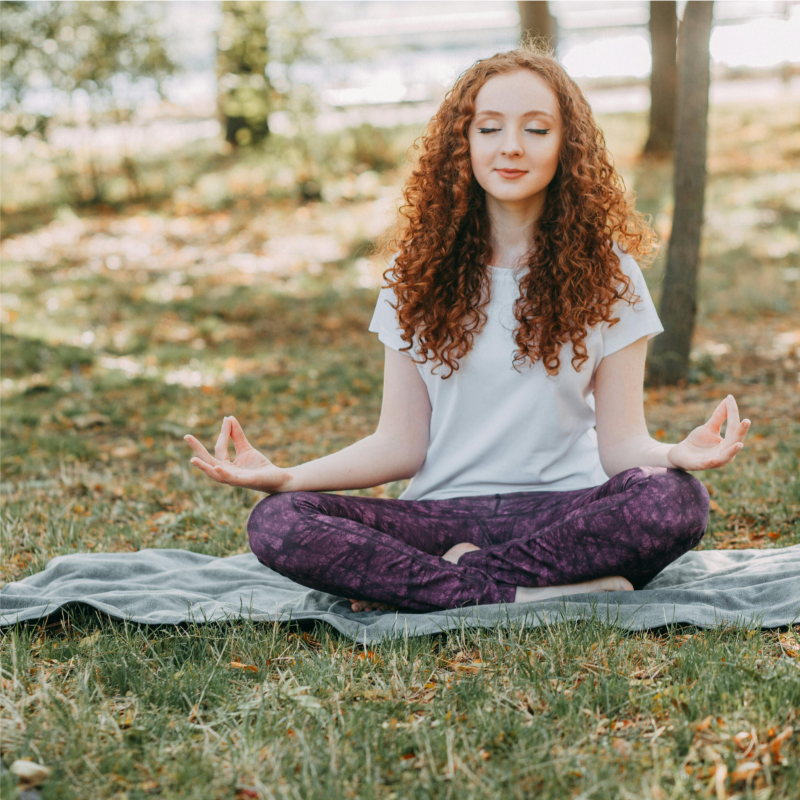 A woman with curly red hair sits cross-legged on a blanket in a park, meditating with her eyes closed and her hands in mudra position, as she contemplates the benefits of meditative walking in mid-mountains.