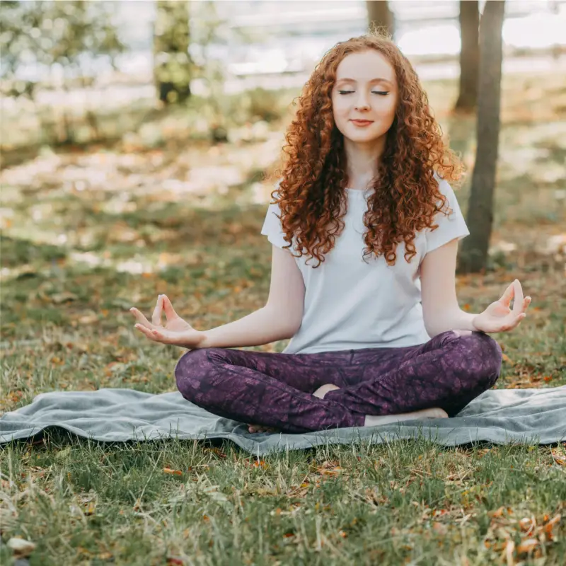 Une femme aux cheveux roux bouclés est assise les jambes croisées sur une couverture dans un parc, méditant les yeux fermés et les mains en position mudra, tandis qu'elle contemple les bienfaits de la marche méditative en moyenne montagne.